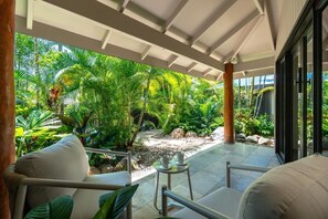 A covered outdoor area with a seating arrangement, a small table, and a view of a tropical garden. - NALA Resort (Mission Beach)