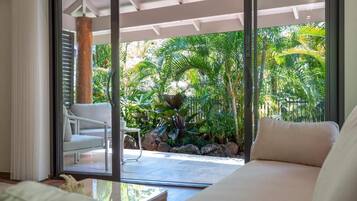 A room with a sliding glass door leading to a patio with a white chair, a sofa, and tropical plants.