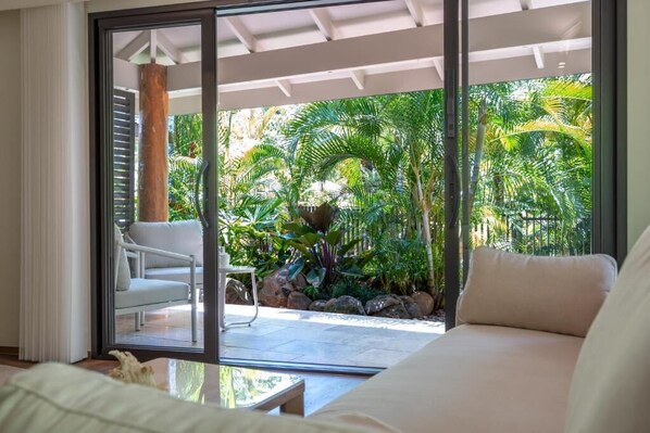 Una habitación con puerta corrediza de vidrio que da a un patio con una silla blanca, un sofá y plantas tropicales.