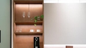 A modern kitchen with wooden cabinetry, a built-in coffee maker, and a chair with a cushioned seat. - NALA Resort (Mission Beach)