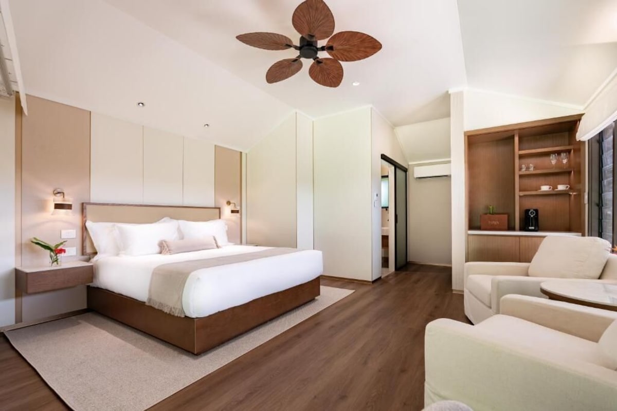 A modern bedroom with a large bed, a ceiling fan, a bedside table with a lamp, and a built-in shelving unit.