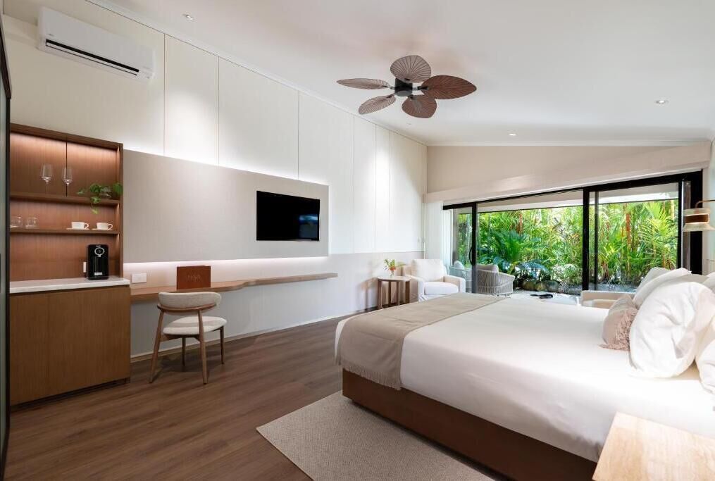 A modern bedroom with a bed, a desk, a chair, a TV, and a view of greenery.