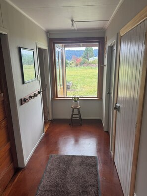 A hallway with wooden flooring, a small stool with a plant, a door with a handle, and a window with a view of a grassy area. - Cosy cottage with beautiful mountain view. Close to town and Carrot Park. (Ohakune)