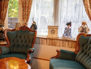 Ein Zimmer mit zwei grünen Polstersesseln, einem Holztisch, einer Uhr und einem Fenster mit Spitzenvorhängen.
