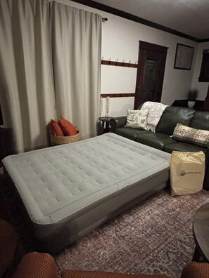 A room with a mattress, a green sofa, a small table, a wicker basket, and a bag on the floor.