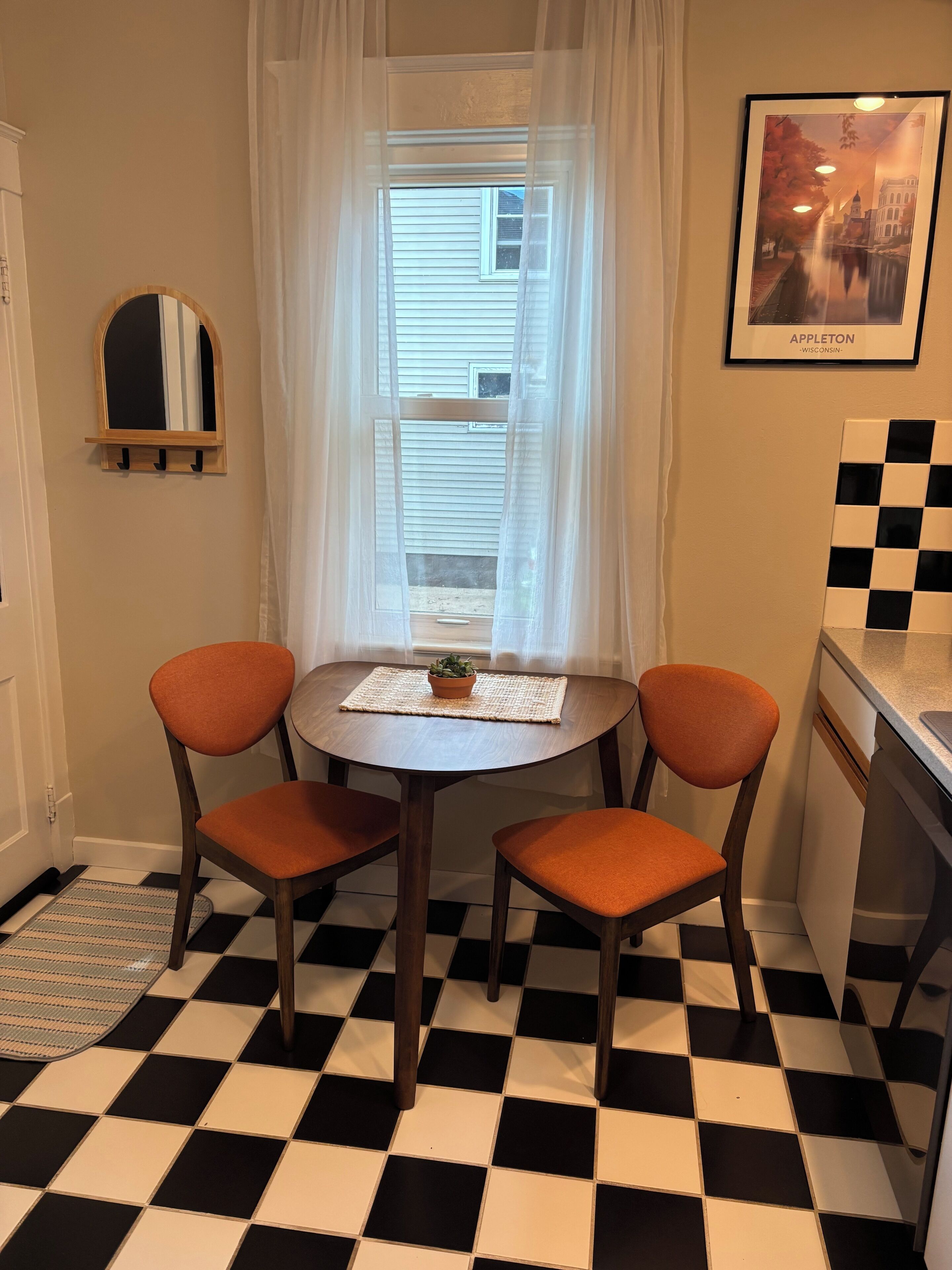 A room with a checkered floor, a round table with two chairs, a window with sheer curtains, a framed picture on the wall, and a small potted plant on the table.