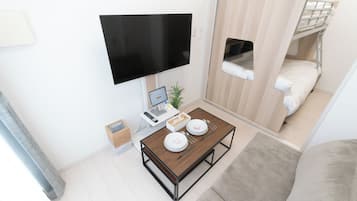 A compact hotel room with a flat-screen TV, a small desk with a computer, a dining table set for two, a bunk bed, and a sofa.