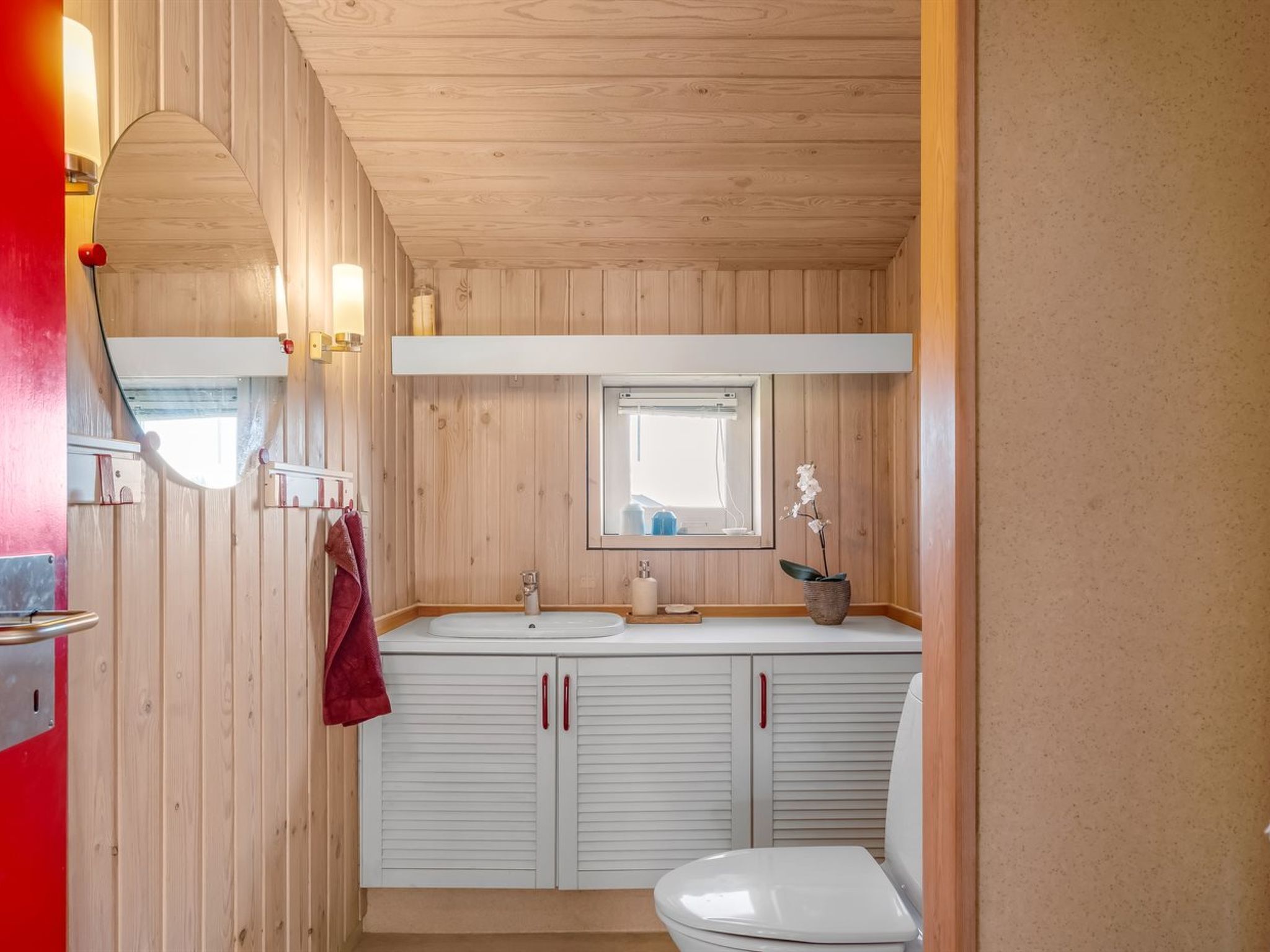 Un baño con paneles de madera, un lavabo blanco, un espejo, un inodoro y una toalla roja colgada en la pared.
