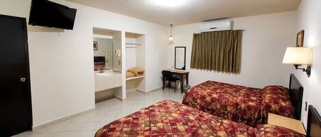 A hotel room with a bed, a desk, a chair, a television, a wardrobe, and an air conditioning unit.