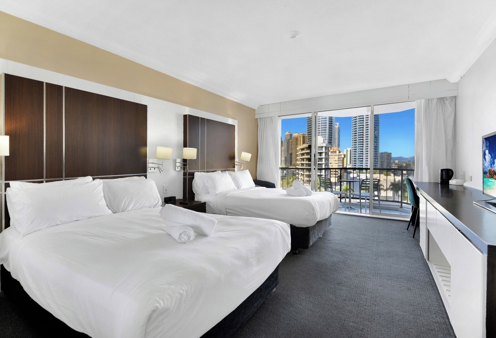 A hotel room with two beds, a desk, a chair, and a balcony with a city view.
