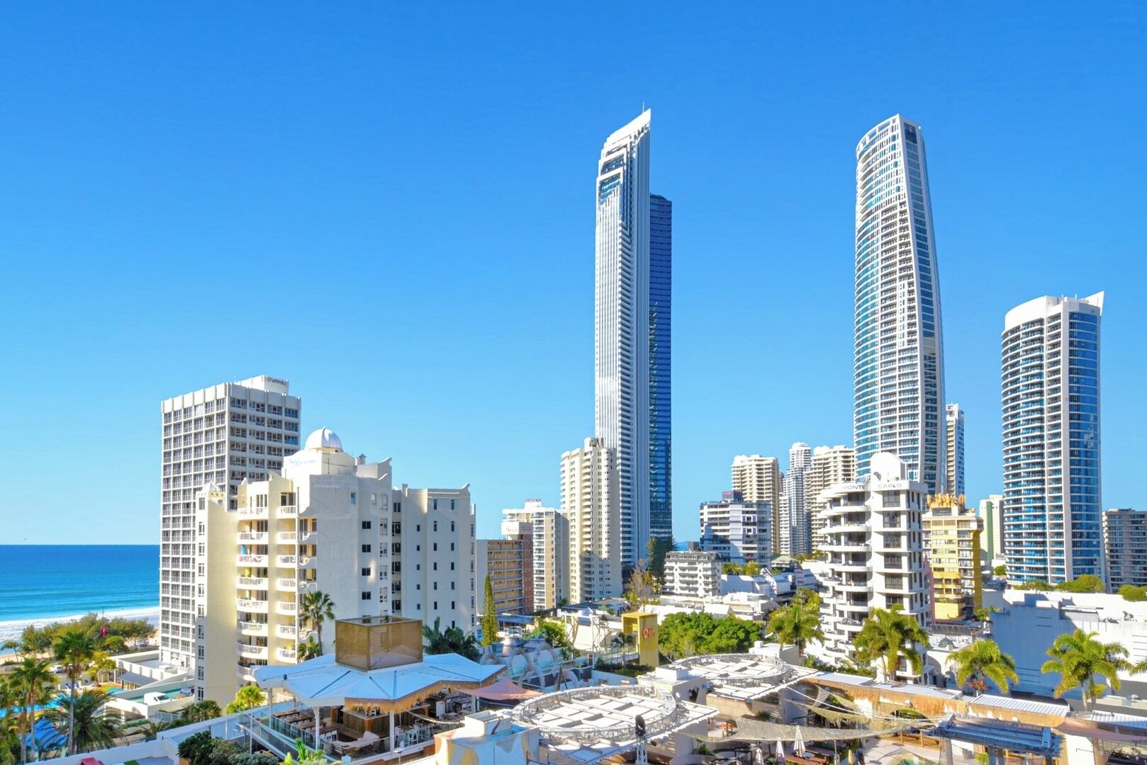 A coastal cityscape with high-rise buildings, a clear blue sky, and a beach in the foreground.