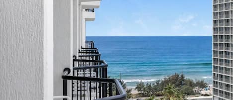 Un balcon avec vue sur l’océan et la plage, un escalier et un grand immeuble en arrière-plan.
