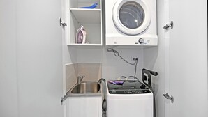 A compact laundry room with a front-loading washer and dryer, a small sink, and a shelf with a blue container and an iron. - Casino-Side 2BR Ocean View | Walk to Beach/Tram (Broadbeach)