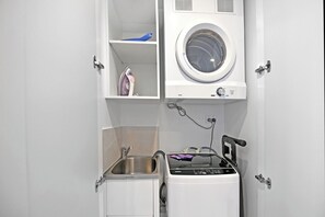 A compact laundry room with a front-loading washer and dryer, a small sink, and a shelf with a blue container and an iron. - Casino-Side 2BR Ocean View | Walk to Beach/Tram (Broadbeach)