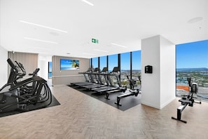 A modern gym with cardio equipment, a TV, and a city view. - Casino-Side 2BR Ocean View | Walk to Beach/Tram (Broadbeach)