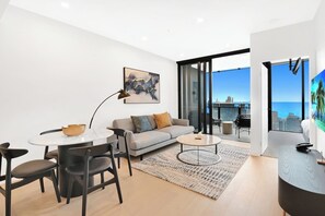 A modern living room with a dining area, a sofa, a coffee table, and a view of the ocean through large windows. - Casino-Side 2BR Ocean View | Walk to Beach/Tram (Broadbeach)