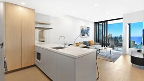 A modern kitchen with a central island, a dining area with a round table, and a living space with a sofa and a city view. - Casino-Side 2BR Ocean View | Walk to Beach/Tram (Broadbeach)
