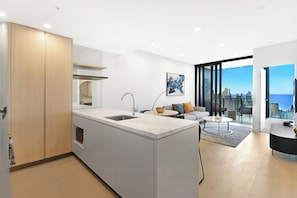 A modern kitchen with a central island, a dining area with a round table, and a living space with a sofa and a city view. - Casino-Side 2BR Ocean View | Walk to Beach/Tram (Broadbeach)