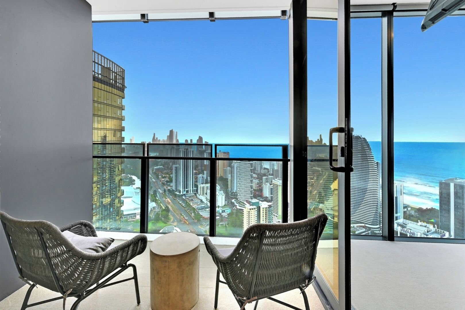 A balcony with two wicker chairs and a small round table, overlooking a cityscape and the ocean.