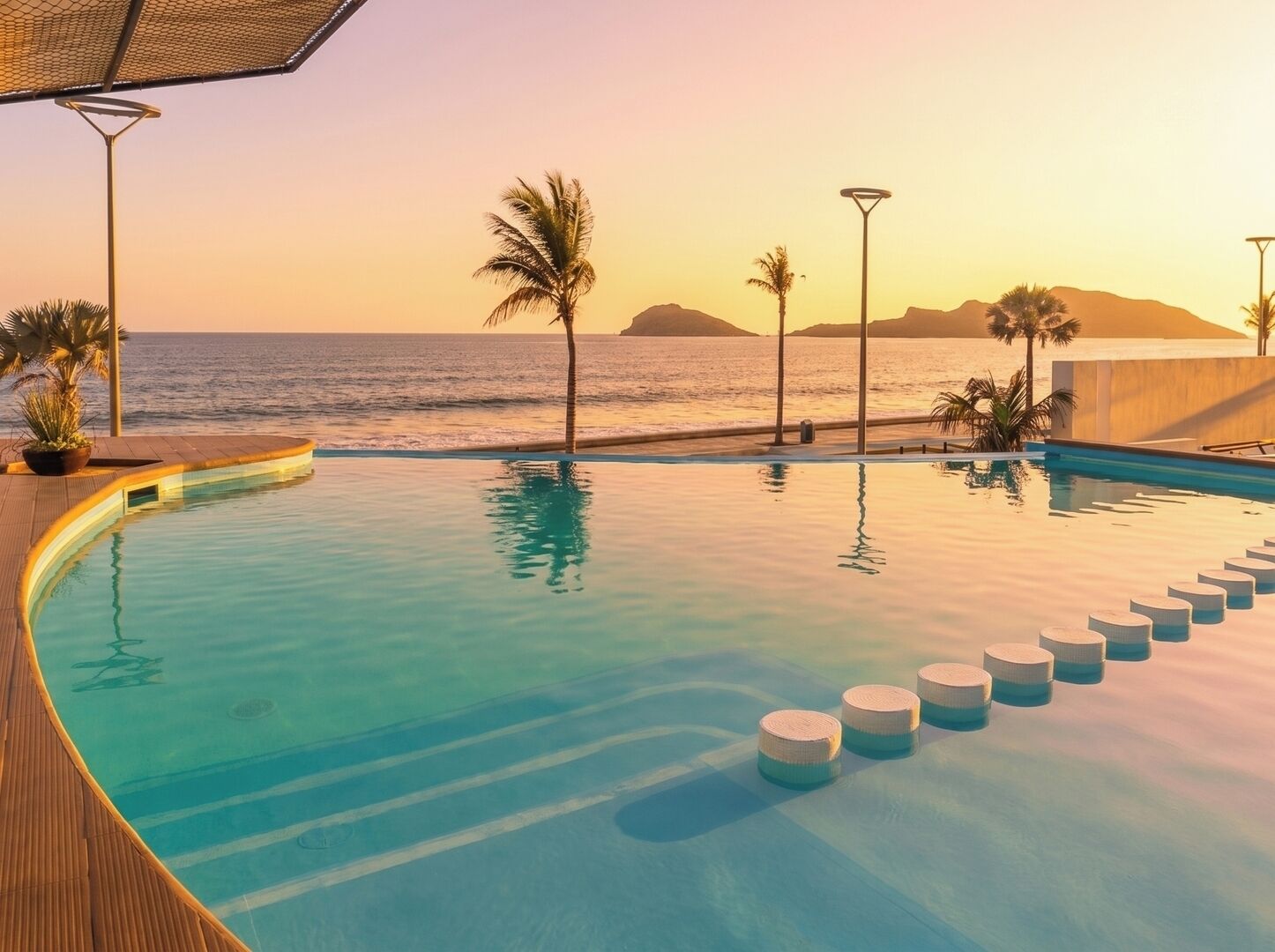 A pool with a view of the ocean at sunset, surrounded by palm trees and a wooden deck.