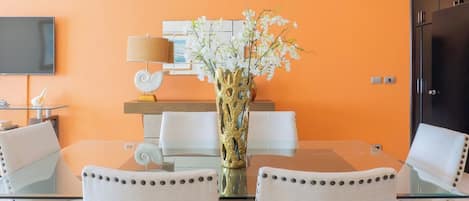 A modern dining area with a glass table, white chairs, a vase with white flowers, a flat-screen TV, and an air conditioning unit on the wall.