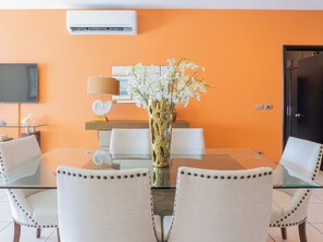 Un espace repas moderne avec une table en verre, des chaises blanches, un vase avec des fleurs blanches, un téléviseur à écran plat et un climatiseur mural.