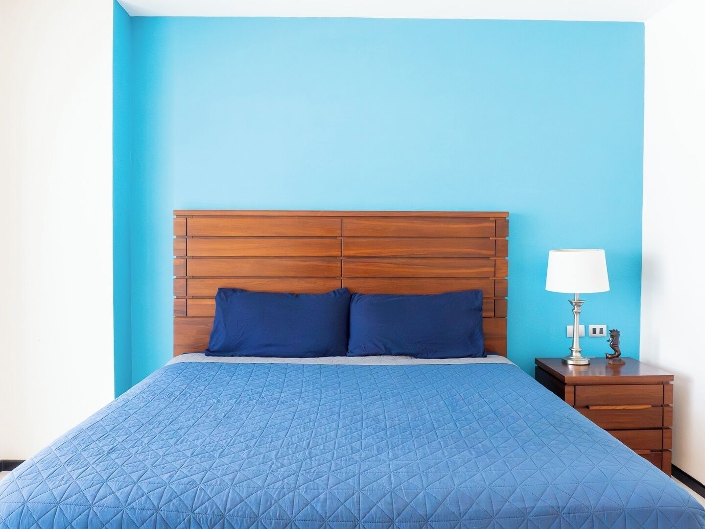 Una cama con cabecera de madera, colcha acolchada azul, dos almohadas, una mesita de noche con lámpara y un pequeño adorno.