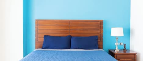Una cama con cabecera de madera, colcha acolchada azul, dos almohadas, una mesita de noche con lámpara y un pequeño adorno.