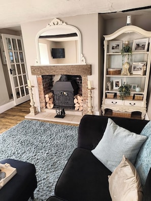 A living room with a fireplace, a black sofa, a blue rug, and a white cabinet with framed photos. - 2BR Cottage for up to 2 adults, 2 children and dogs (Wiltshire)