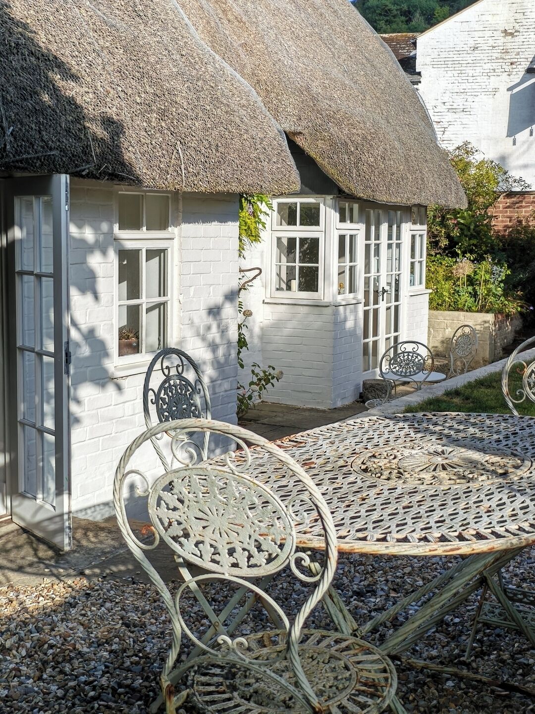 Une petite maison blanche au toit de chaume, avec une porte blanche et une fenêtre à cadre blanc. Devant la maison, on trouve une table en métal accompagnée d’une chaise en métal.