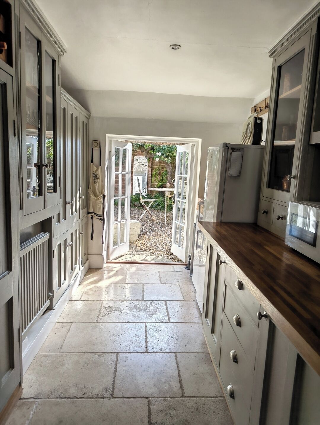 A kitchen with open doors leading to a garden, stone flooring, and wooden cabinets.