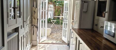 A kitchen with open doors leading to a garden, stone flooring, and wooden cabinets.