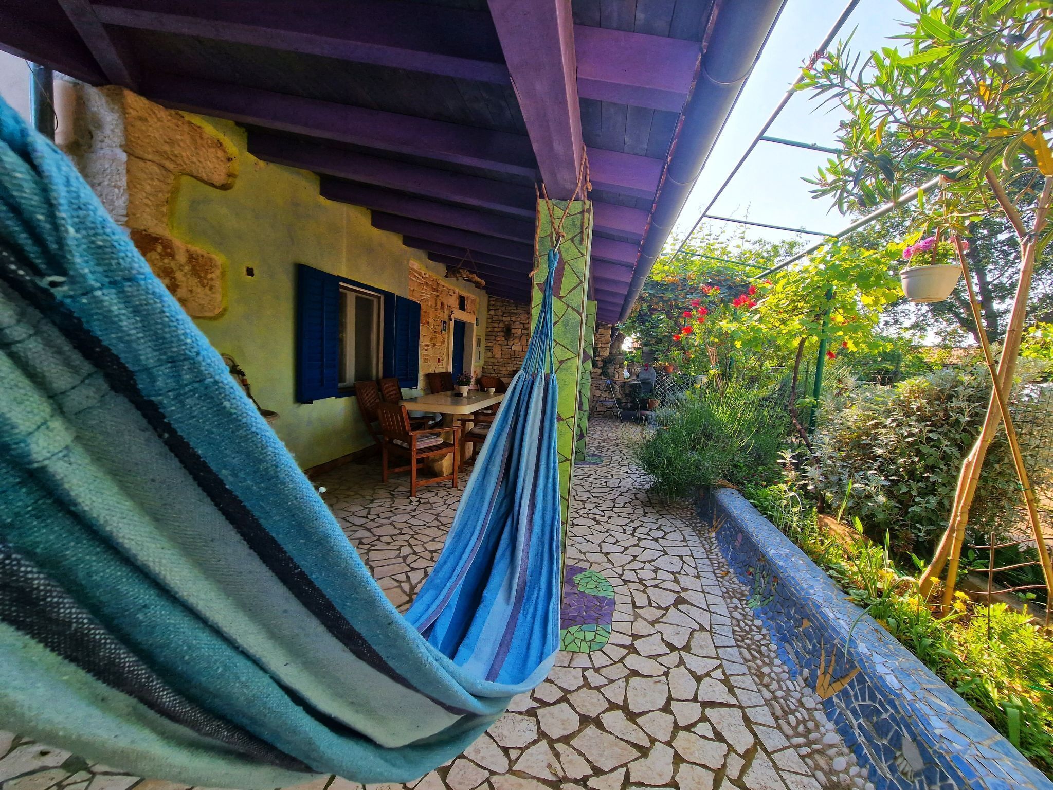 Eine Hängematte unter einer überdachten Terrasse, eine terrasse mit Steinplatten und ein rustikales Gebäude mit Tür und Fenster.