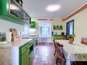 A kitchen with green cabinets, a dining table with chairs, a window with lace curtains, and a shelf with jars. - Vacation home Helena by Interhome (Pula)