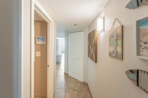 A hallway with a white door, a tiled floor, and wall-mounted artwork. - 2BR Oceanfront View at Carolinian with Amazing  Pool (Myrtle Beach)