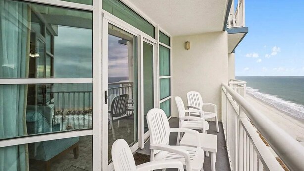 A balcony with white chairs, a table, and a view of the ocean. - 2BR Oceanfront View at Carolinian with Amazing  Pool (Myrtle Beach)