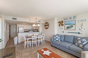 A modern living room with a sofa, a dining table with chairs, a kitchen area, and a wall with nautical decor. - 2BR Oceanfront View at Carolinian with Amazing  Pool (Myrtle Beach)