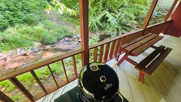 A barbecue grill on a wooden deck with a bench, overlooking a stream and dense greenery.