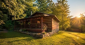 A log cabin with a porch, surrounded by a stone wall and a grassy yard, set in a wooded area.