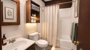 A bathroom with a white sink, toilet, and bathtub. There is a wooden shelf above the toilet with towels and toiletries.