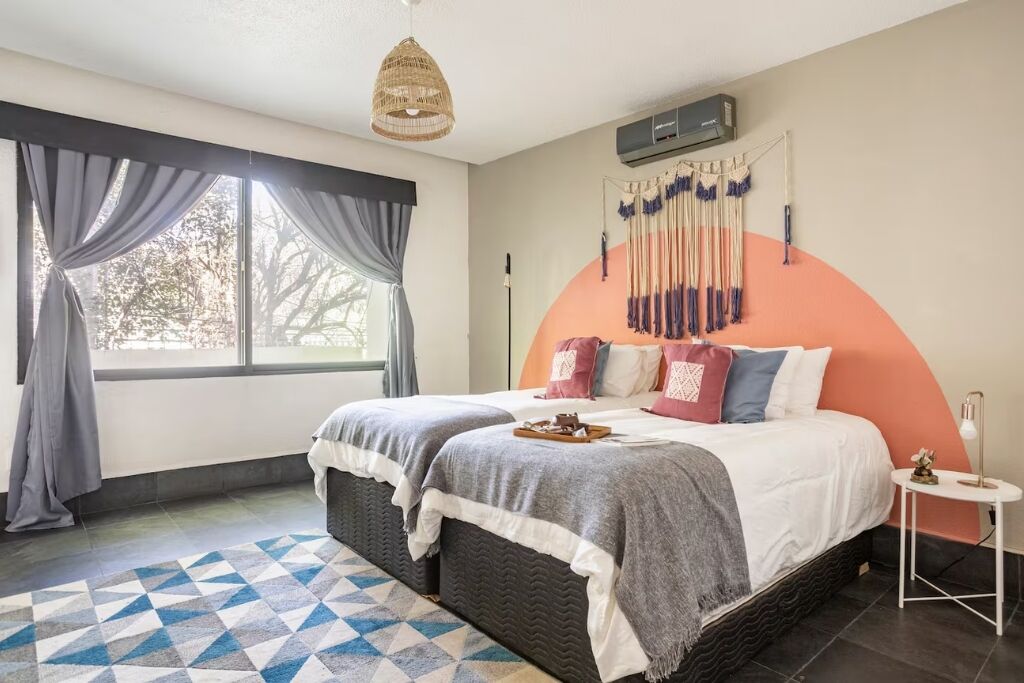 A bedroom with a large bed, a hanging light fixture, a wall-mounted air conditioner, and a view through a window with curtains.