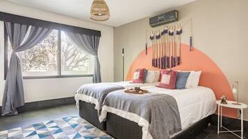 A bedroom with a large bed, a hanging light fixture, a wall-mounted air conditioner, and a view through a window with curtains.