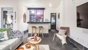A modern living room with a sofa, coffee table, and bar stools. There is a kitchen area with a large window and a TV mounted on the wall.