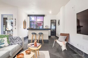 A modern living room with a sofa, coffee table, and bar stools. There is a kitchen area with a large window and a TV mounted on the wall. - Large Private Condesa Home • Perfect for Families & Groups (Ciudad de México)