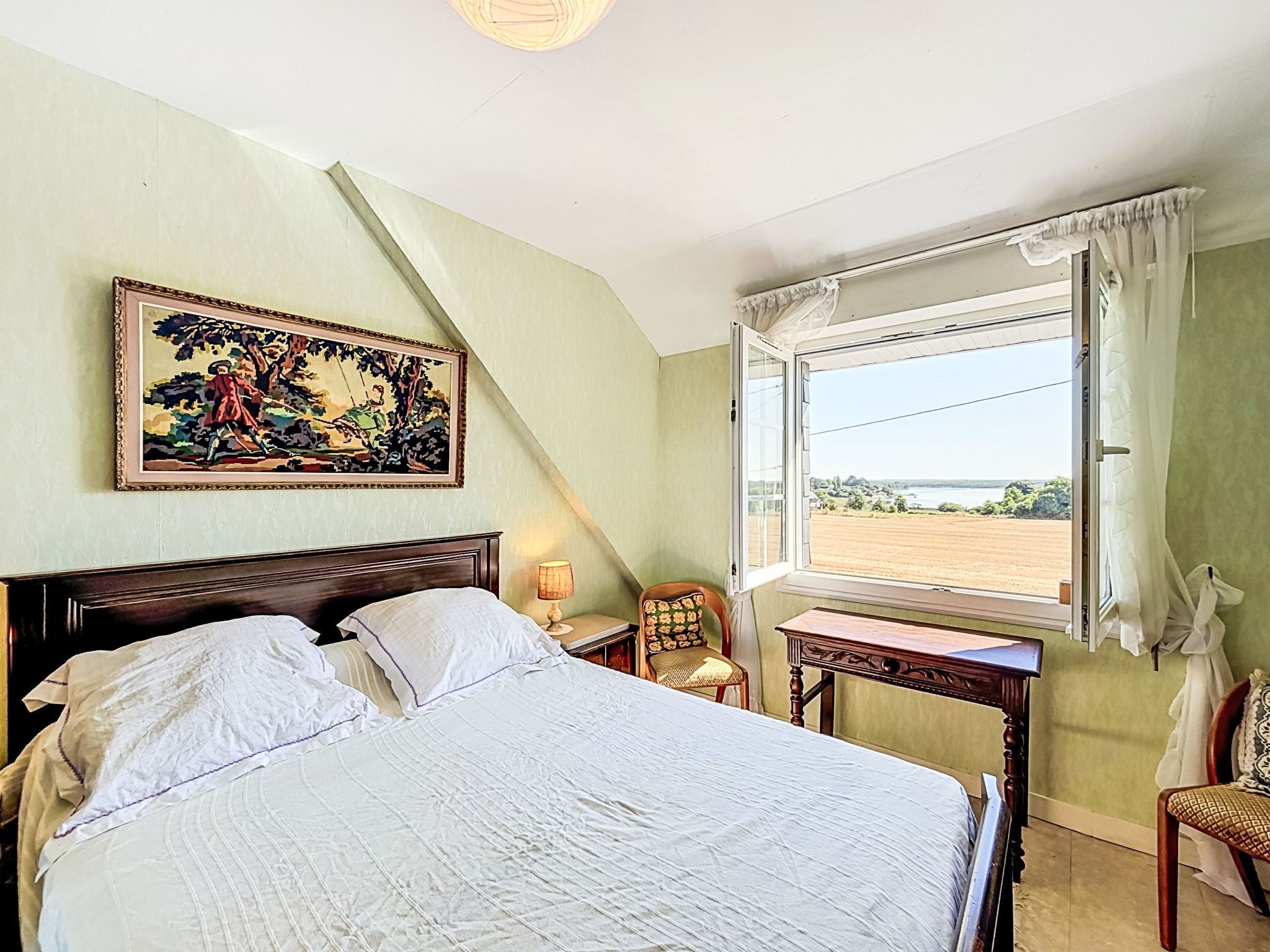 Une chambre à coucher avec un lit, une chaise, une table, une fenêtre avec des rideaux et un tableau au mur.