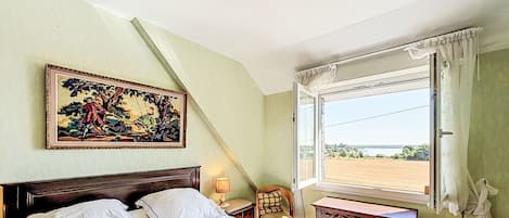 Un dormitorio con cama, una silla, una mesa, una ventana con cortinas y un cuadro en la pared.