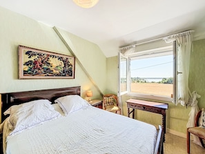 Ein Schlafzimmer mit Bett, Stuhl, Tisch, einem Fenster mit Vorhängen und einem Bild an der Wand.