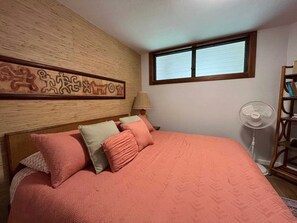 A bedroom with a bed, pillows, a wall-mounted artwork, a window, a standing fan, and a bookshelf. - Oceanfront SeaVillage Penthouse 1 bed 1 bath near Kona Village downtown (Kailua-Kona)