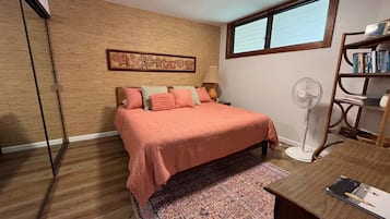 Un dormitorio con cama, escritorio, estantería, ventilador y ventana.