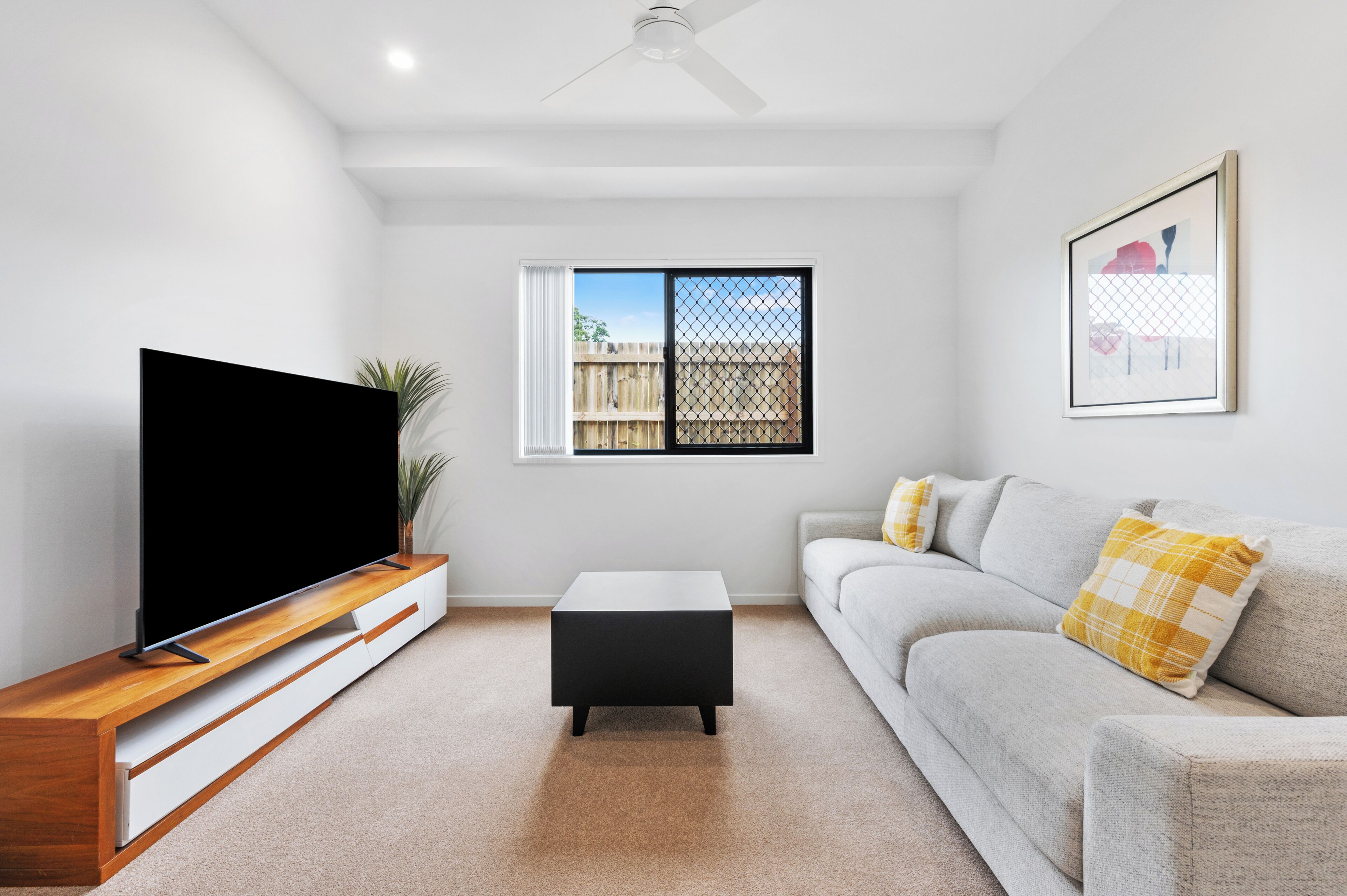 A living room with a flat-screen TV, a coffee table, a sofa, a ceiling fan, and a window with a view of a fence.